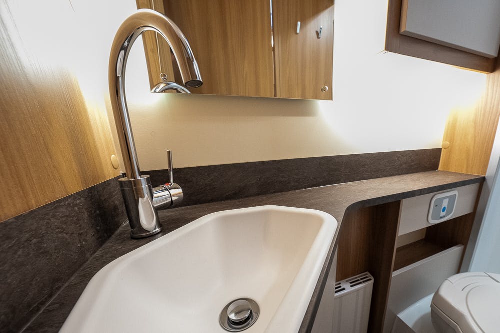 A modern bathroom countertop in the 2022 Bailey Autograph 74-4 features a white sink with an elegant silver faucet. The countertop is made of dark material, and there is a mirrored cabinet above it. The walls and furnishings are wooden and beige tones. A toilet is partially visible in the background.