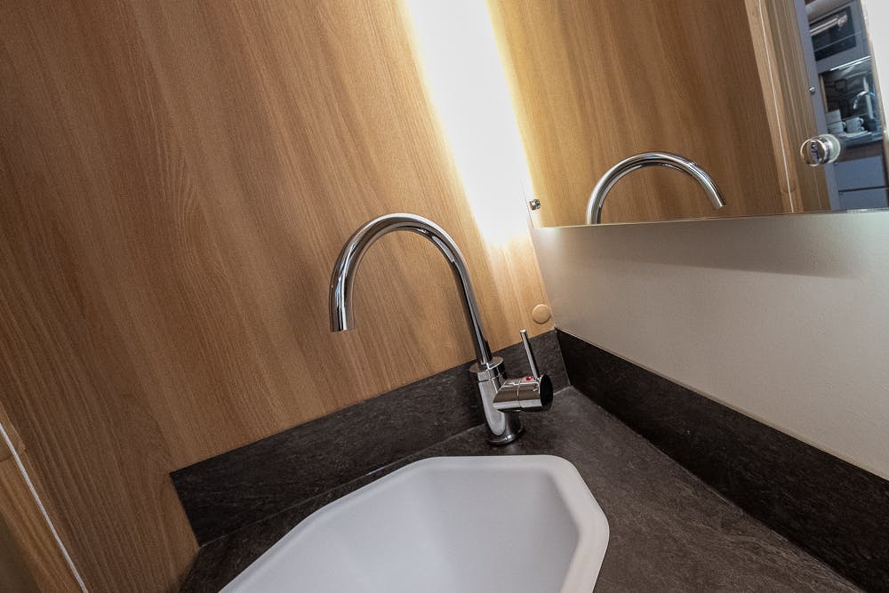 A modern bathroom sink with a tall, curved faucet mounted on a dark countertop is shown. A wooden backsplash and a large mirror are visible behind the sink. The area is well-lit by an overhead light, embodying the sleek design found in the 2022 Bailey Autograph 74-4 RVs.