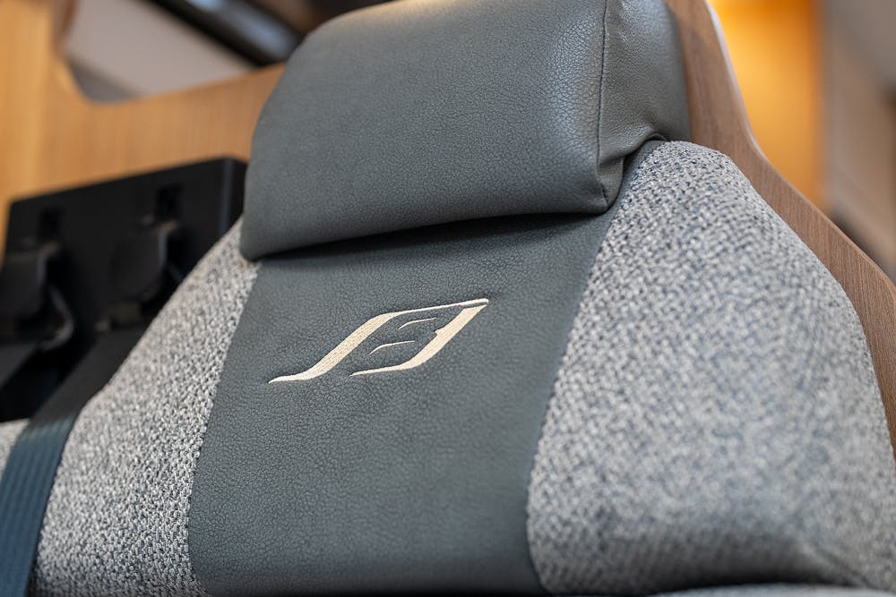 Close-up of a car seat headrest in the 2022 Bailey Autograph 74-4, featuring a logo with a stylized leaf. The headrest is covered in gray leather and gray fabric near the edges. The background includes part of a wooden panel and some out-of-focus elements.