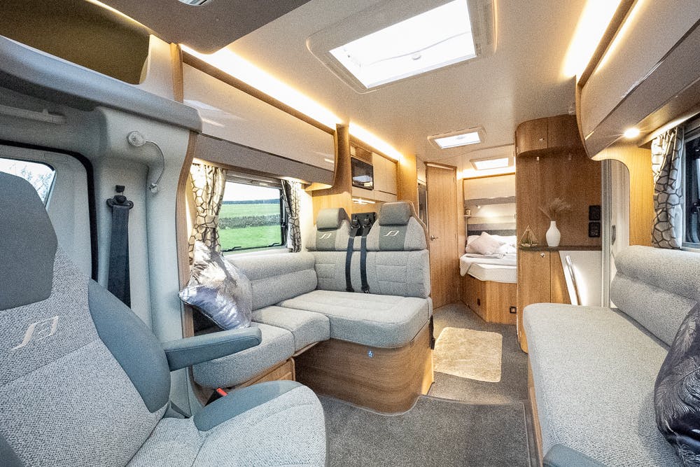 A modern, well-lit 2022 Bailey Autograph 74-4 interior featuring a cozy seating area with grey upholstered seats, a small bed in the background, overhead storage compartments, and large windows with patterned curtains. The space is clean and neatly organized.
