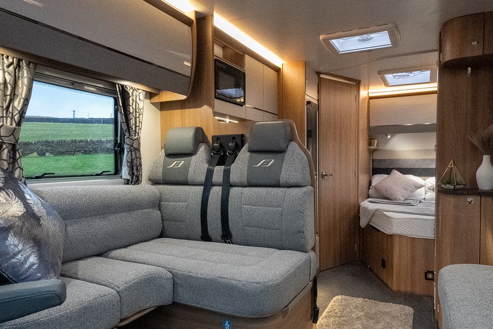 Interior of the 2022 Bailey Autograph 74-4 motorhome featuring a cozy seating area with gray upholstered seats, a window with patterned curtains, and ample natural light. The bedroom area, with a neatly made bed, is visible in the background. Warm, wooden tones dominate the decor.