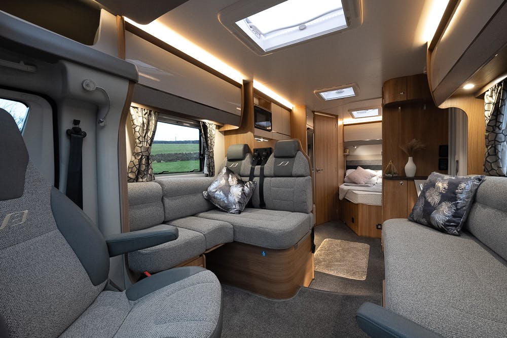 Interior of a modern 2022 Bailey Autograph 74-4 RV with grey upholstered seating, throw pillows, wooden cabinetry, and a small bed in the background. Skylights illuminate the space, and windows with blackout curtains are visible. The floor is carpeted, adding to the cozy atmosphere.