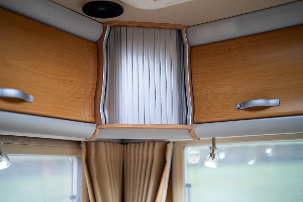 The image displays the interior of a 2007 Auto-Sleepers Sigma EL with wooden cabinets and a vertical metal sliding door. Below the cabinets, there is a window with cream-colored curtains and two small ceiling spotlights providing light.