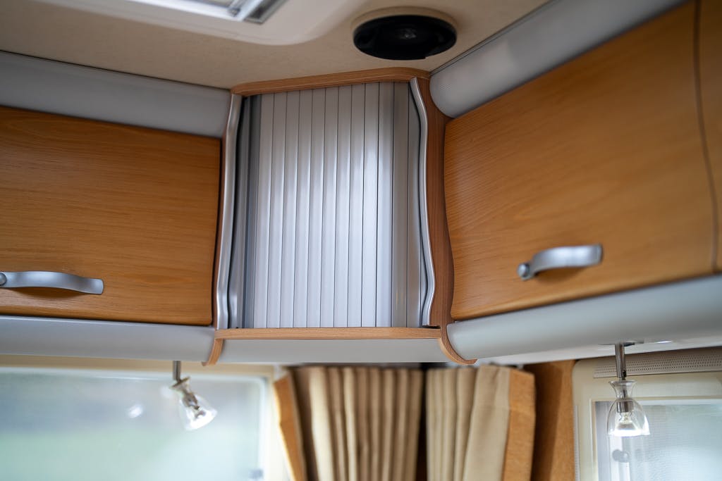 The image shows the interior of a 2007 Auto-Sleepers Sigma EL camper van. It features wooden cabinets with silver handles. In the center, there is a vertical tambour door made of silver slats. Below, beige curtains frame a window with metal rail lights on either side.