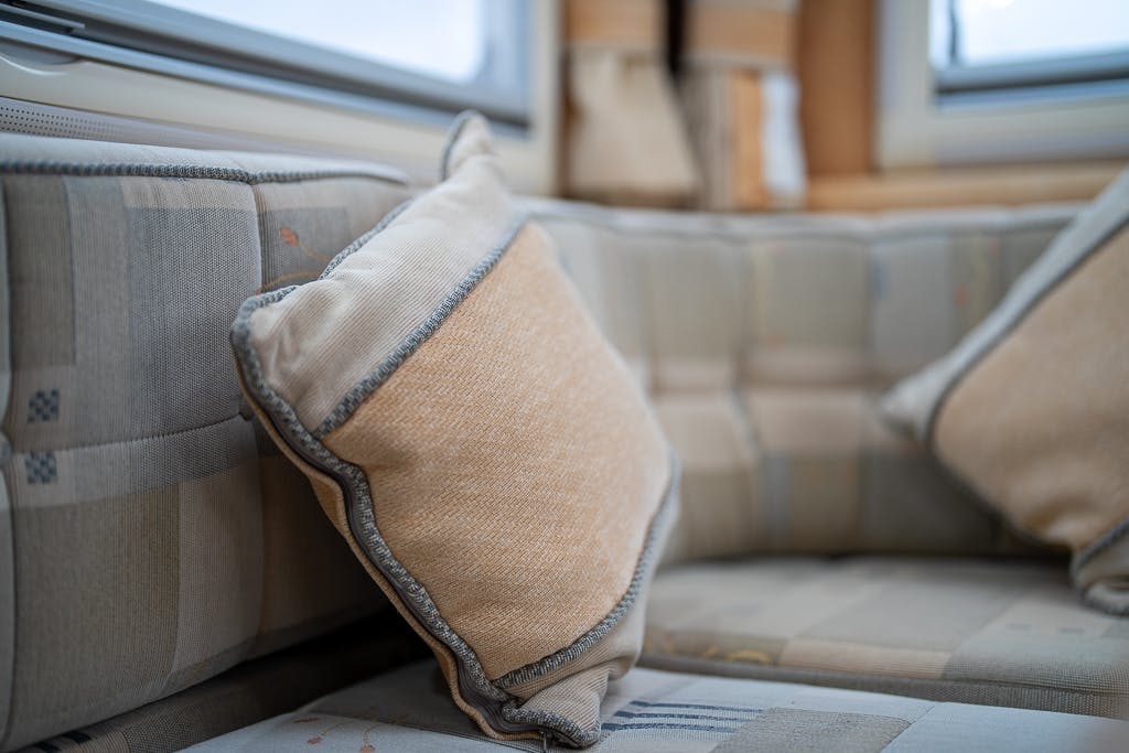 A close-up shot shows a beige throw pillow leaning against a cushioned sofa. The upholstery, reminiscent of the 2007 Auto-Sleepers Sigma EL, features a subtle checkered pattern. The background includes a window with beige curtains, slightly out of focus.