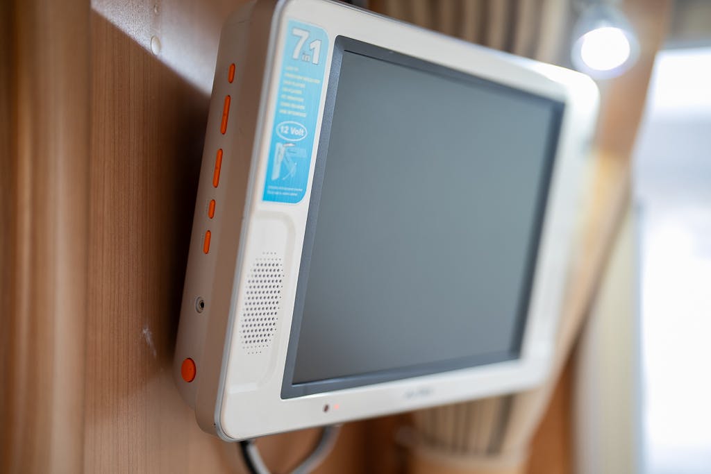 A flat screen monitor is mounted on a wooden wall in the 2007 Auto-Sleepers Sigma EL. The monitor has a white frame with several orange buttons on the side and a blue sticker on the top-left corner indicating 7.1 and 12 volts. The screen is blank.