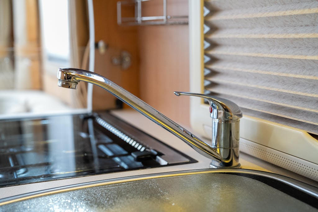A modern kitchen faucet with a single lever handle is installed on a countertop next to a black stove, reminiscent of the sophistication seen in 2007 Auto-Sleepers Sigma EL. The background features a window with beige blinds and wooden cabinetry, complementing the shiny chrome finish of the faucet.