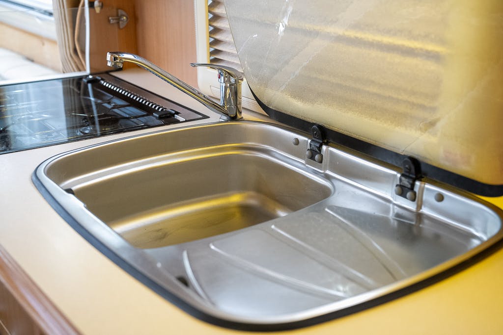 The image shows a standard stainless steel kitchen sink with a single basin installed in the countertop of a 2007 Auto-Sleepers Sigma EL. The sink has a faucet to the left and a hinged lid that covers part of it when closed. The beige countertop is next to a stovetop.