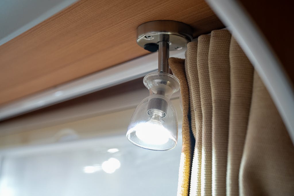 A small, wall-mounted reading light is switched on in the cozy interior of a 2007 Auto-Sleepers Sigma EL. The light has a clear glass shade and is attached to a wooden panel above a beige curtain. The background is partially blurred, showing another light reflection on a window.