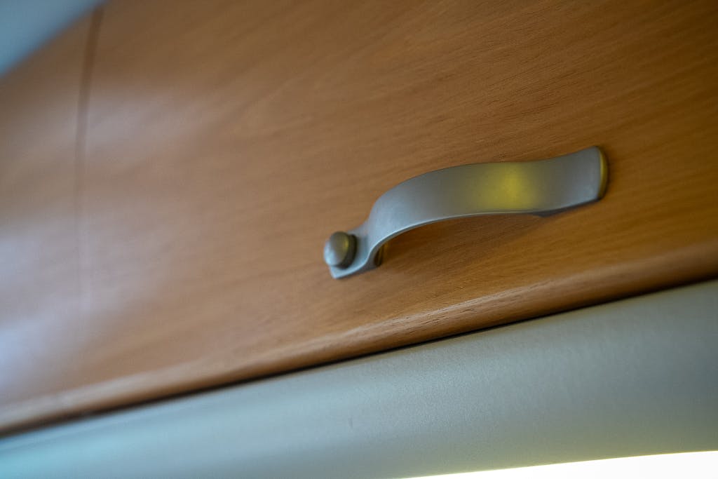 Close-up view of a wooden cabinet door with a metal handle in the 2007 Auto-Sleepers Sigma EL. The handle has a sleek, curved design with a small round attachment point. The wood has a light, warm tone.