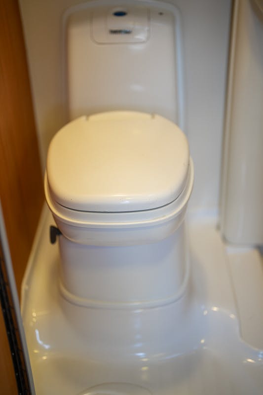 A compact white plastic toilet with a closed lid installed in a small, presumably mobile space such as a 2007 Auto-Sleepers Sigma EL camper or boat. The interior walls and floor are also white, and a small control panel is visible at the back.