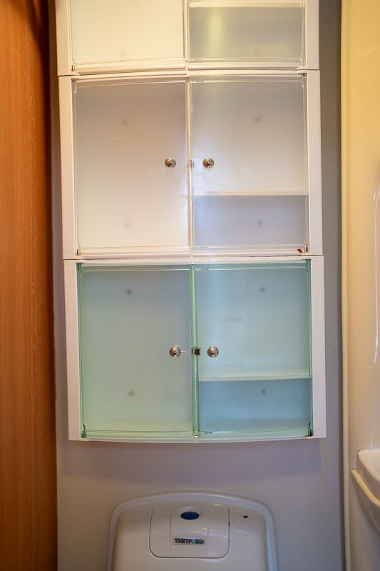 A wall-mounted bathroom cabinet with frosted glass doors and metal knobs is shown. The cabinet consists of four compartments, two with glass panels and two empty. Below the cabinet is a toilet with a flush button on top, reminiscent of the sleek design found in the 2007 Auto-Sleepers Sigma EL.