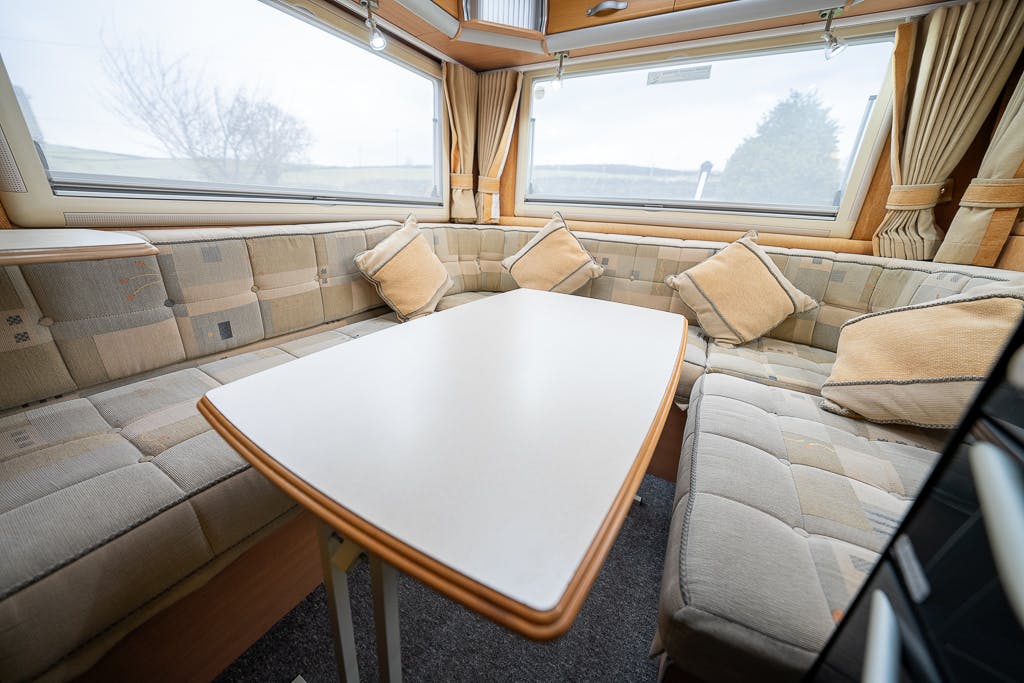 The interior of the 2007 Auto-Sleepers Sigma EL caravan features a spacious seating area with beige cushioned seats arranged in a U-shape around a rectangular table. Several matching pillows are placed on the seats, and large windows provide natural light and outdoor views.