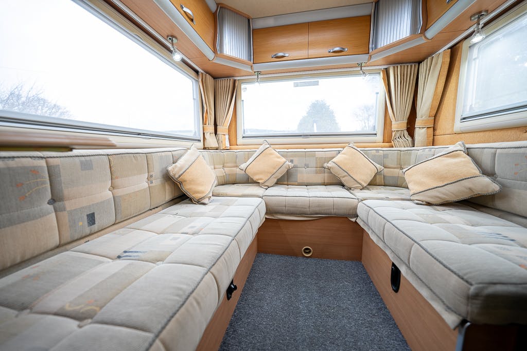 The interior of the 2007 Auto-Sleepers Sigma EL camper van features an inviting L-shaped seating area with beige, cushioned upholstery. The space includes matching pillows and windows with curtains on both sides, allowing ample natural light to flood in.