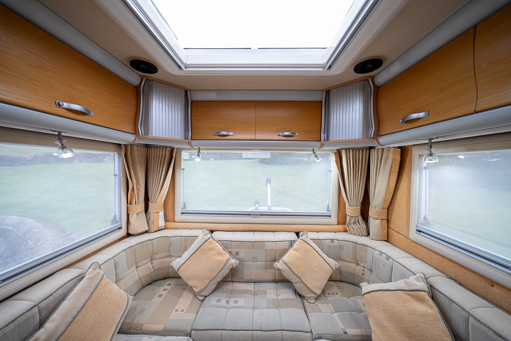 Interior of a cozy 2007 Auto-Sleepers Sigma EL camper van showing a U-shaped cushioned seating area with patterned upholstery, two windows with pulled-back curtains, overhead storage cabinets, and a skylight above. Several throw pillows are placed on the seating.