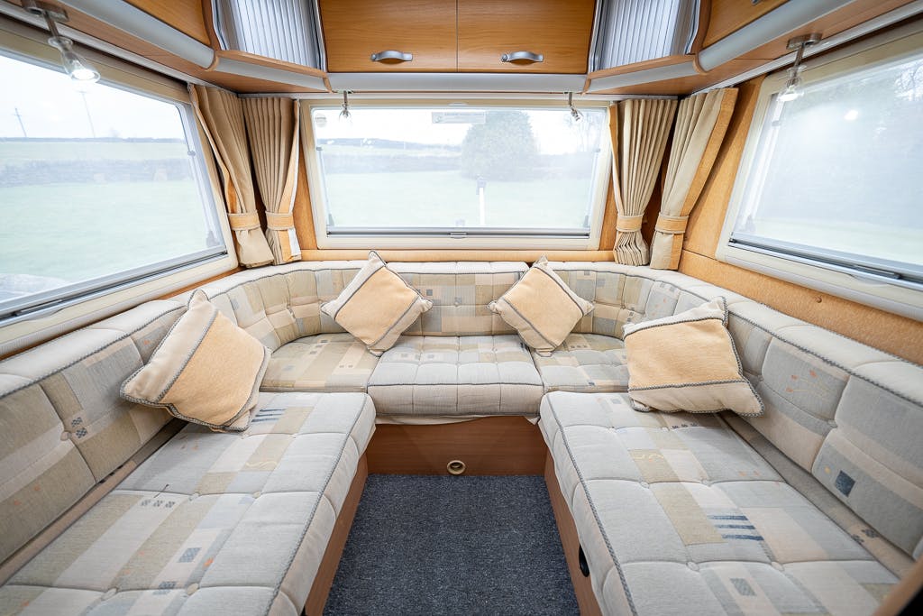 The image shows the interior of a 2007 Auto-Sleepers Sigma EL caravan, featuring an L-shaped seating arrangement with beige patterned cushions. The seating area is surrounded by large windows with beige curtains, allowing for ample natural light.