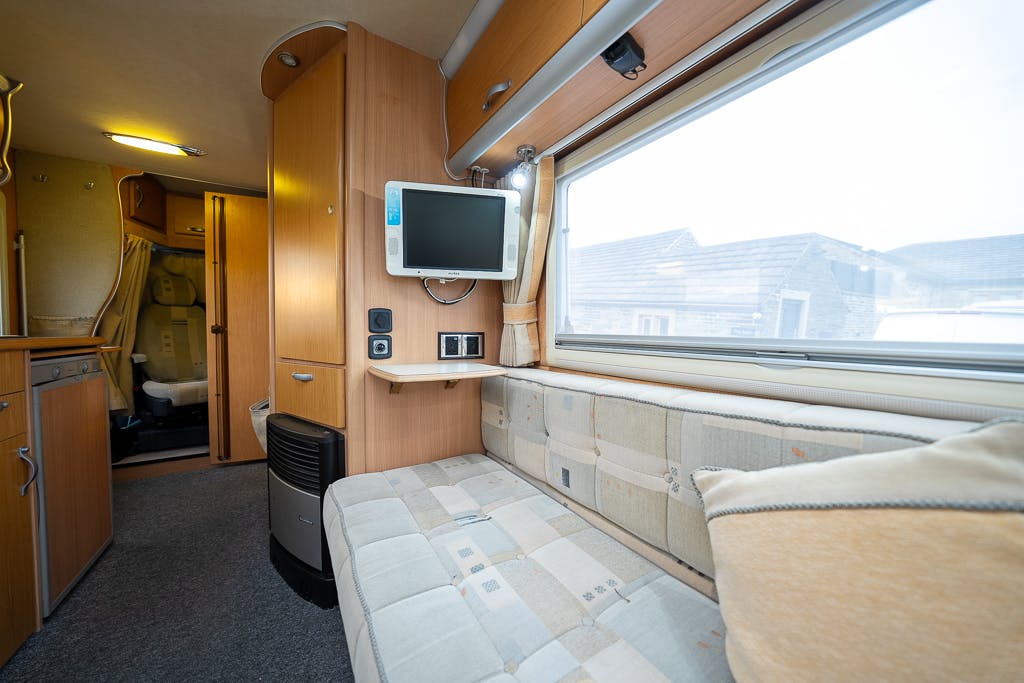 The image shows the interior of a 2007 Auto-Sleepers Sigma EL camper van. There is a cushioned bench with a pillow next to a large window. Above the bench is a flat-screen TV mounted on the wooden wall. A kitchenette and bathroom are visible towards the back of the van.