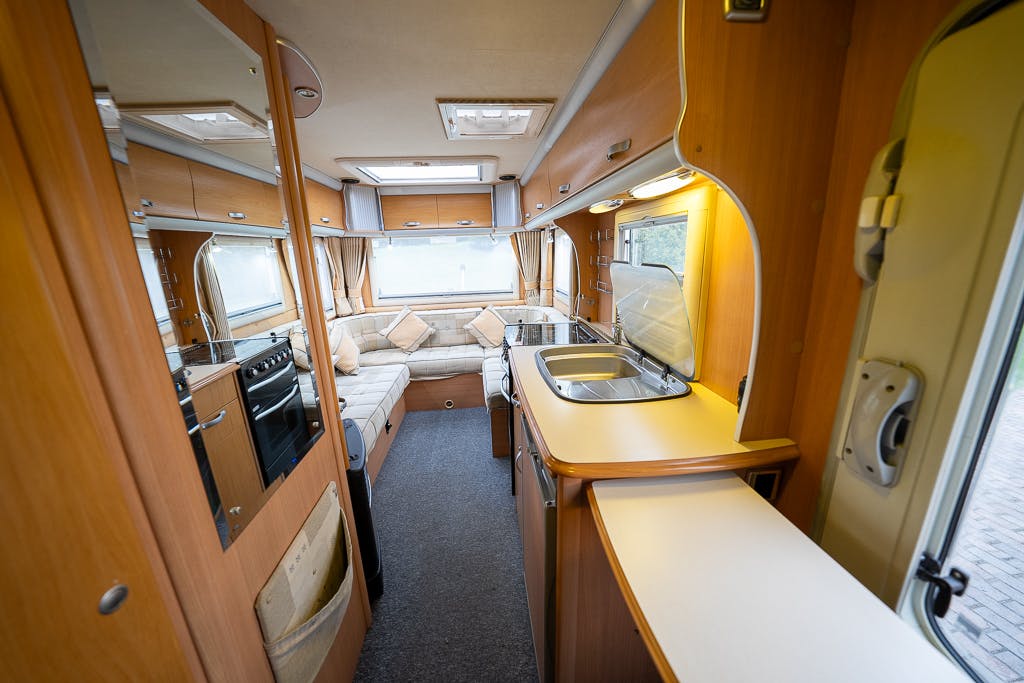 Interior of a parked 2007 Auto-Sleepers Sigma EL RV with wooden cabinets and grey carpet. The modern kitchen area includes a stove and sink on the right. The back features a large, cushion-filled seating area with windows. Bright overhead lights and a skylight provide illumination.