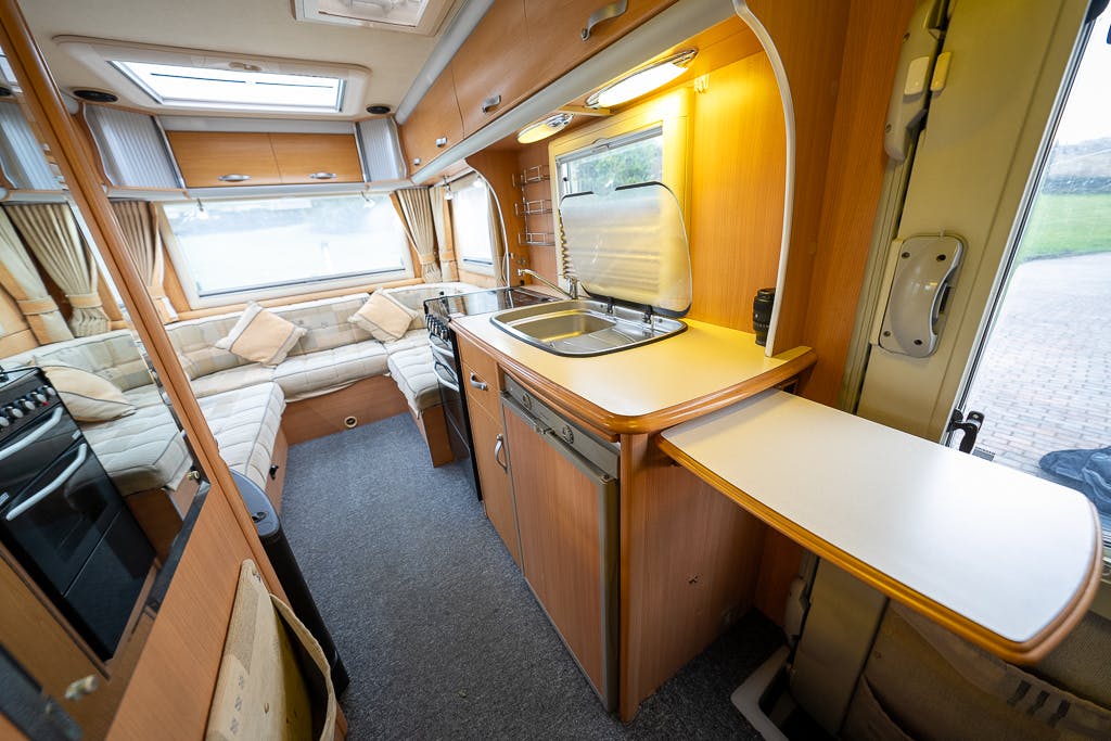Interior of a 2007 Auto-Sleepers Sigma EL featuring a compact kitchen with a sink, stove, and countertop space. Adjacent to the kitchen is a seating area with a U-shaped sofa and a large window. The decor primarily consists of wood paneling with beige accents.