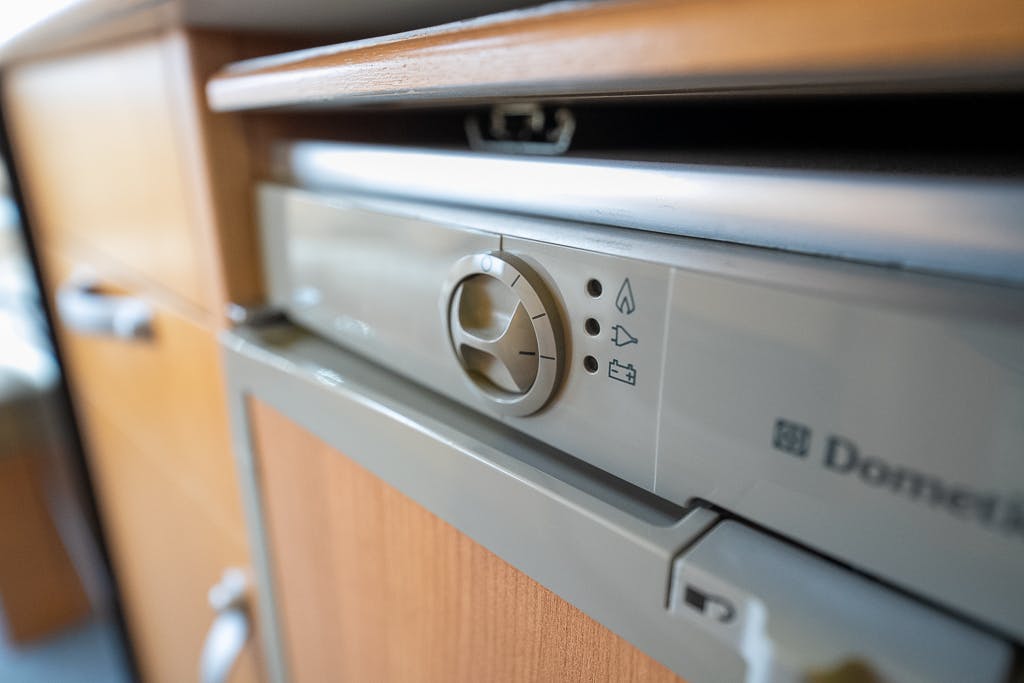 Close-up view of a control panel on a kitchen appliance, featuring a rotary knob and three symbols indicating various functions. The beige and wooden-finish unit, reminiscent of the 2007 Auto-Sleepers Sigma EL design, has part of a countertop visible above.
