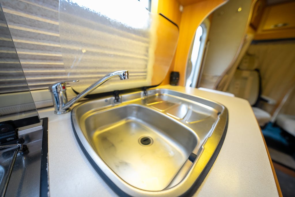 A close-up of a stainless steel sink in a 2007 Auto-Sleepers Sigma EL camper van kitchen. The sink has a curved faucet and is set into a clean, white countertop. The background shows the interior of the camper, including a window with a blind and seating area.