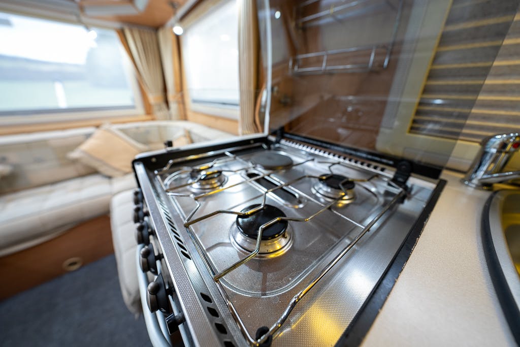 The image shows a four-burner gas stove inside a 2007 Auto-Sleepers Sigma EL camper van. The stove is built into the countertop and has a glass cover that is propped open. Behind the stove, there is a seating area with a beige cushion and a window with curtains.
