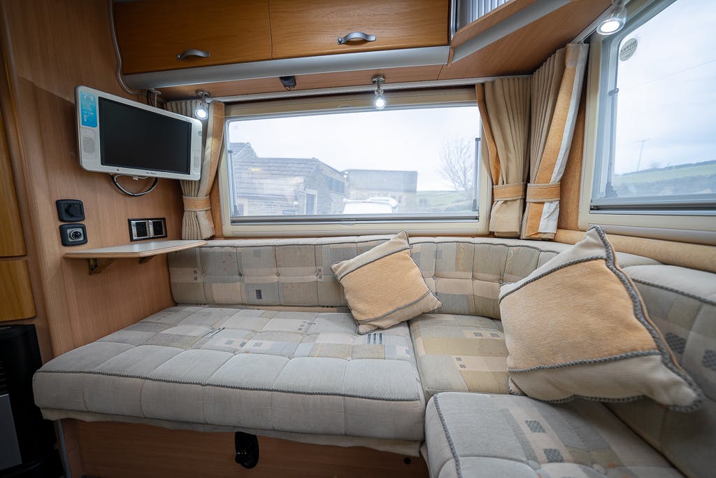 The interior of the 2007 Auto-Sleepers Sigma EL features a corner seating area with a cushioned L-shaped bench, beige and grey patterned upholstery, and matching pillows. Above the seating area is a window with beige curtains. A small TV is mounted on the left wall.