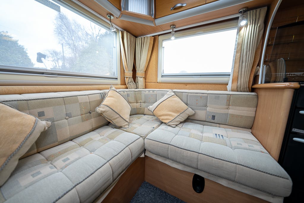 Interior of a 2007 Auto-Sleepers Sigma EL camper van with a corner seating area. The seating is upholstered in light-colored fabric with a plaid pattern and comes with matching cushions. Windows with curtains provide natural light, and cabinetry is visible in the background.