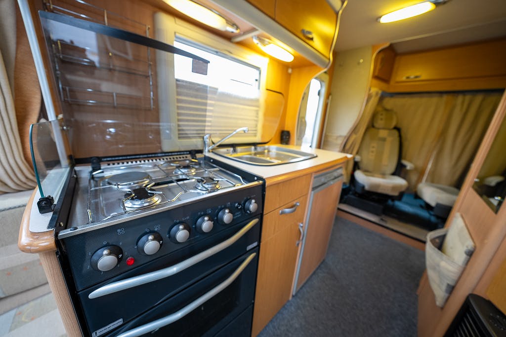 The image shows the interior of a 2007 Auto-Sleepers Sigma EL campervan kitchen area, featuring a stovetop with four burners, an oven, a sink, and wooden cabinets. The stove has control knobs on the front. Beyond the kitchen, a driver's seat and passenger seat are visible.