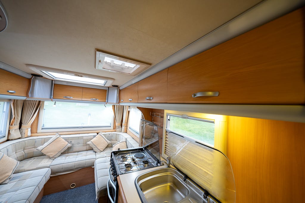 Interior of a 2007 Auto-Sleepers Sigma EL caravan showing a kitchen area with a gas stove, dual-basin sink, and wooden cabinets. Adjacent is a seating area with cushioned benches and large windows. The space is well-lit and neatly organized.