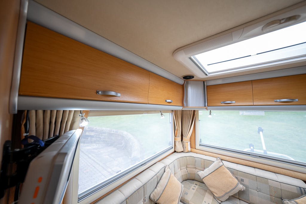 The image shows the interior of a 2007 Auto-Sleepers Sigma EL with a view of overhead wooden cabinets with metal handles. Below, there is a U-shaped cushioned seating area with light brown and beige pillows. Windows along the walls provide natural light.