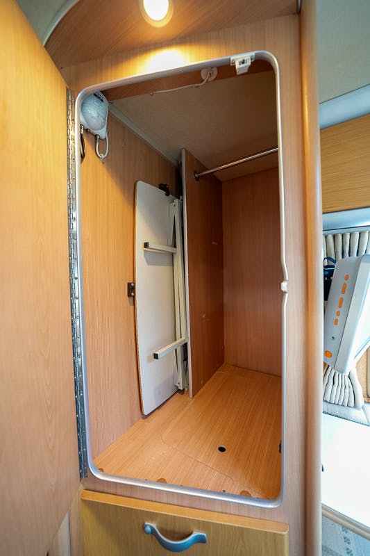 An empty wooden closet with a single shelf, a hanging rod, and a light fixture at the top inside a room. A white folded tabletop or panel is attached to one side of the closet, reminiscent of the clever design seen in the 2007 Auto-Sleepers Sigma EL, and the closet door appears to be open, revealing its interior.
