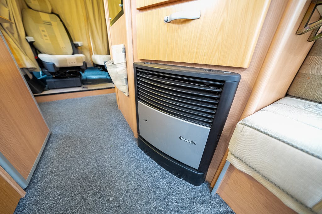 The image shows the interior of a 2007 Auto-Sleepers Sigma EL motorhome, focusing on a heater unit installed at the bottom of a wooden panel. Carpet flooring is visible, with a driver's seat and a beige cushioned seat also in view.