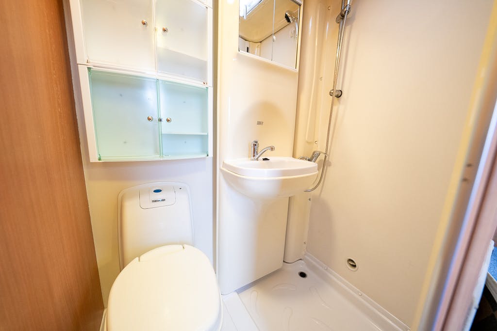A compact bathroom in the 2007 Auto-Sleepers Sigma EL features a toilet, a small sink with a faucet, and a tall showerhead. Above the sink, a mirrored cabinet with frosted glass doors enhances space efficiency. The design boasts minimal fixtures and an all-white color scheme for an airy feel.