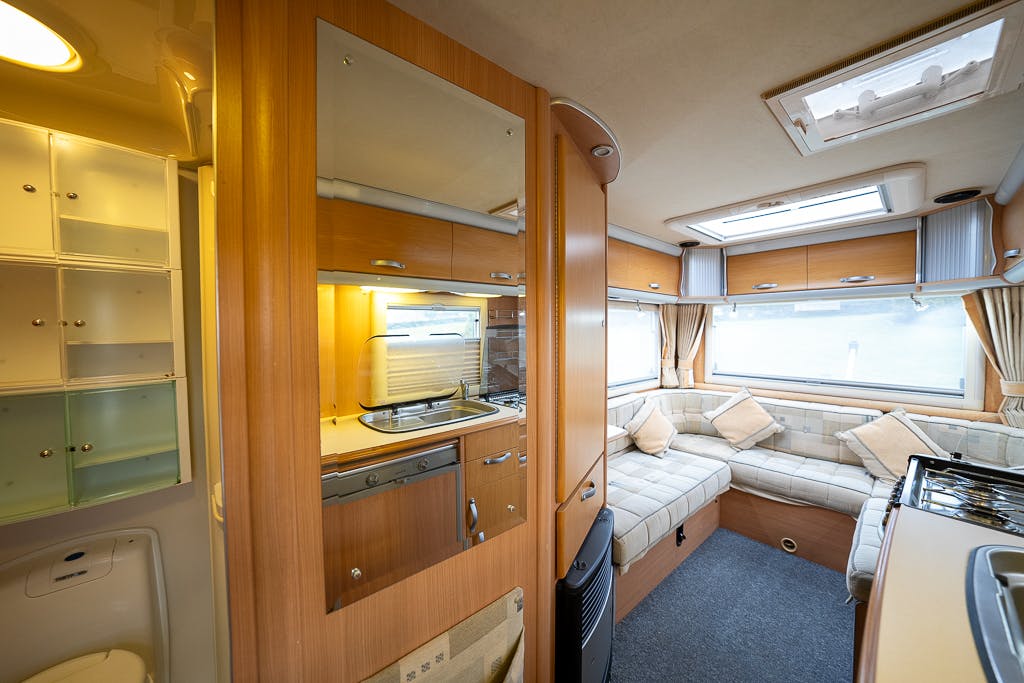 The image shows the interior of a well-lit 2007 Auto-Sleepers Sigma EL camper van. The kitchen area on the left includes a stove, oven, and cabinets. The living area on the right has an L-shaped sofa with cushions. Skylights and windows provide natural light in the space.