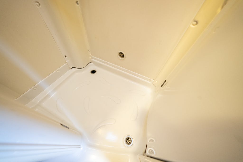 A close-up view of a clean, white shower stall inside a 2007 Auto-Sleepers Sigma EL. The image shows the lower part of the stall, including the curved walls, a round drain in the floor, and a small circular fixture on the back wall. The surface is smooth and shiny.