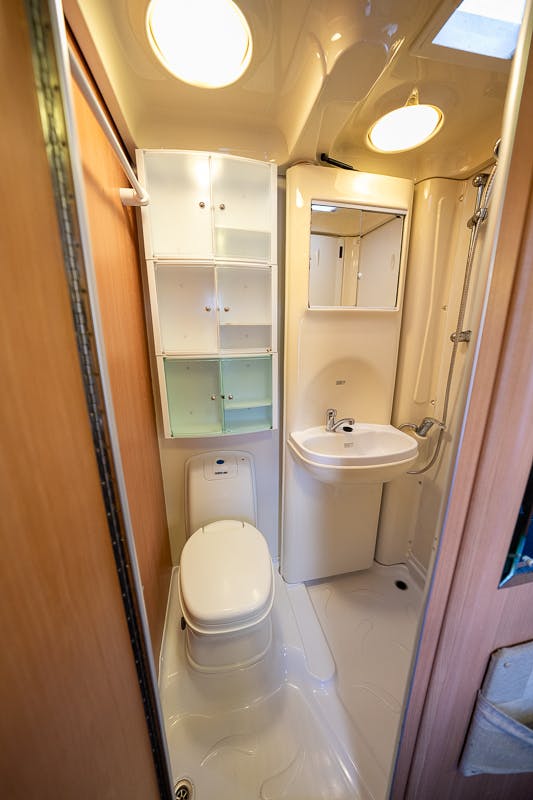 A compact bathroom in the 2007 Auto-Sleepers Sigma EL features a white toilet, sink, and shower area. Above the sink is a mirrored cabinet, and on the left wall are additional white storage shelves. The space boasts light wood finishes and is well-lit with ceiling lights.