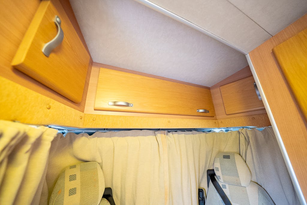 Close-up of the interior of a 2007 Auto-Sleepers Sigma EL motorhome, showcasing the top part of the driver's and passenger's seats, closed overhead storage cabinets, and beige curtains covering windows. The cabinet handles are metallic and the seats feature patterned upholstery.
