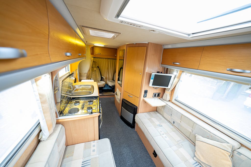 The image shows the interior of a modern 2007 Auto-Sleepers Sigma EL motorhome. It features a compact kitchen area with a sink and stove on the left, bench seating on both sides, a mounted television, and a driver's cab area towards the back. The decor includes light wood cabinetry and neutral-colored upholstery.