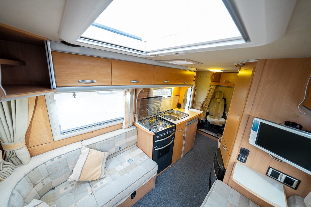 The image shows the interior of a 2007 Auto-Sleepers Sigma EL with light wood cabinetry. Features include a kitchenette with a sink, stovetop, and oven, a small seating area with patterned cushions, and a mounted flat-screen TV. The space is well-lit with natural light from a skylight.
