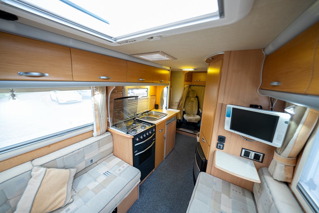The image shows the interior of a 2007 Auto-Sleepers Sigma EL compact RV. The space features a small kitchen with a stove and sink, a built-in seat with storage cabinets overhead, and a wall-mounted TV. Light wood finishes and bright upholstery give it a cozy and organized appearance.