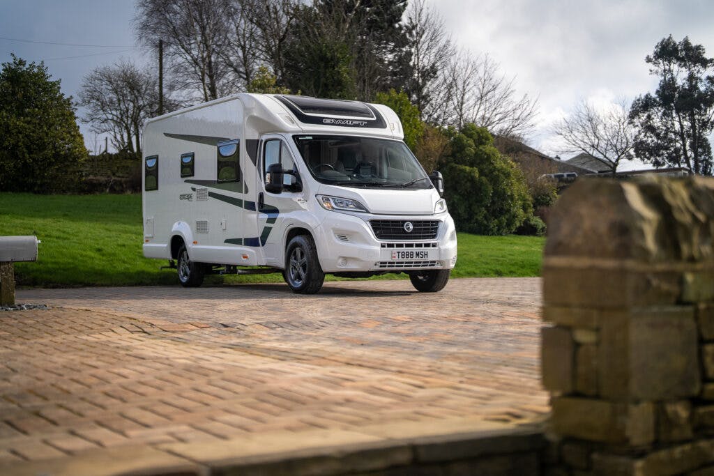 A 2017 Swift Escape 664 motorhome is parked on a brick driveway with grass and trees in the background. The vehicle has a side door, windows, and visible branding on the side. The sky is partly cloudy, and a stone structure is seen in the foreground.