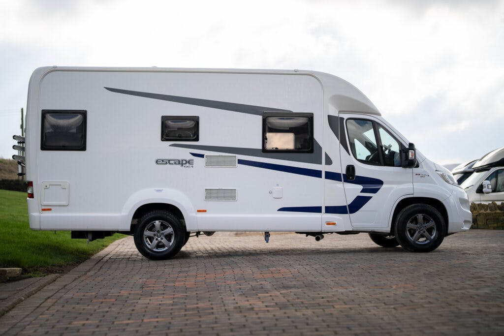 A white 2017 Swift Escape 664 motorhome, with "Escape" written on its side, is parked on a paved area. The motorhome features dark-tinted windows and minimal blue and grey accents. In the background, a grassy area meets a partly cloudy sky.