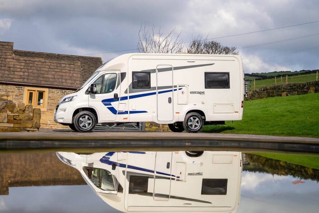 A 2017 Swift Escape 664 motorhome with blue accents is parked on a paved surface, with greenery and a stone building in the background. The vehicle's reflection is visible in a body of water in the foreground, under a cloudy sky.