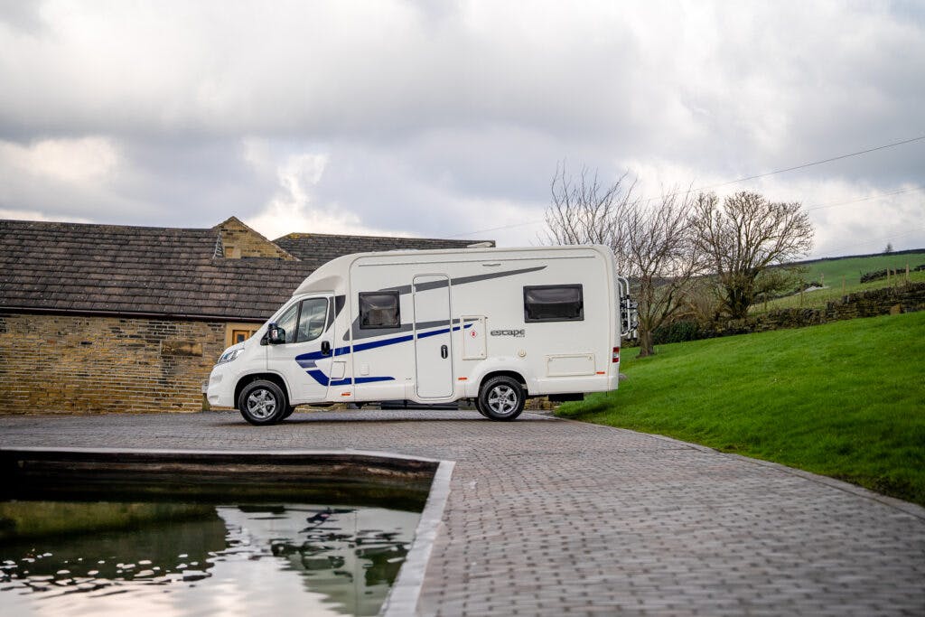 A 2017 Swift Escape 664 motorhome, with blue accents, is parked on a cobblestone driveway next to a reflective body of water. In the background, grassy terrain and a stone building appear under a cloudy sky.