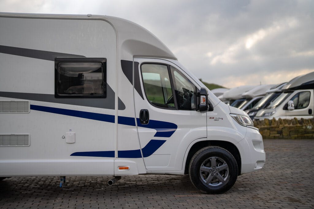 A 2017 Swift Escape 664, a white camper van with blue and gray detailing, is parked on a cobblestone surface. The van features a large side window and has a modern design. More camper vans can be seen in the background under a cloudy sky.