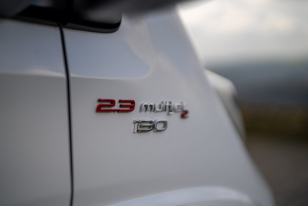 Close-up of a white 2017 Swift Escape 664 showing a badge that reads "2.3 Multijet 130." The background is blurred, making the focus on the vehicle's badge.