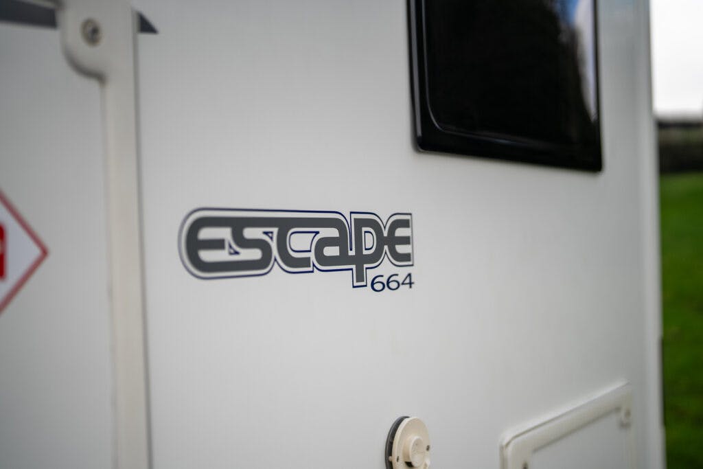 Close-up of a 2017 Swift Escape 664's side showing the logo "Escape 664" in black and gray lettering. A window and a circular component are also visible on the vehicle's surface. The background shows a green grassy area.