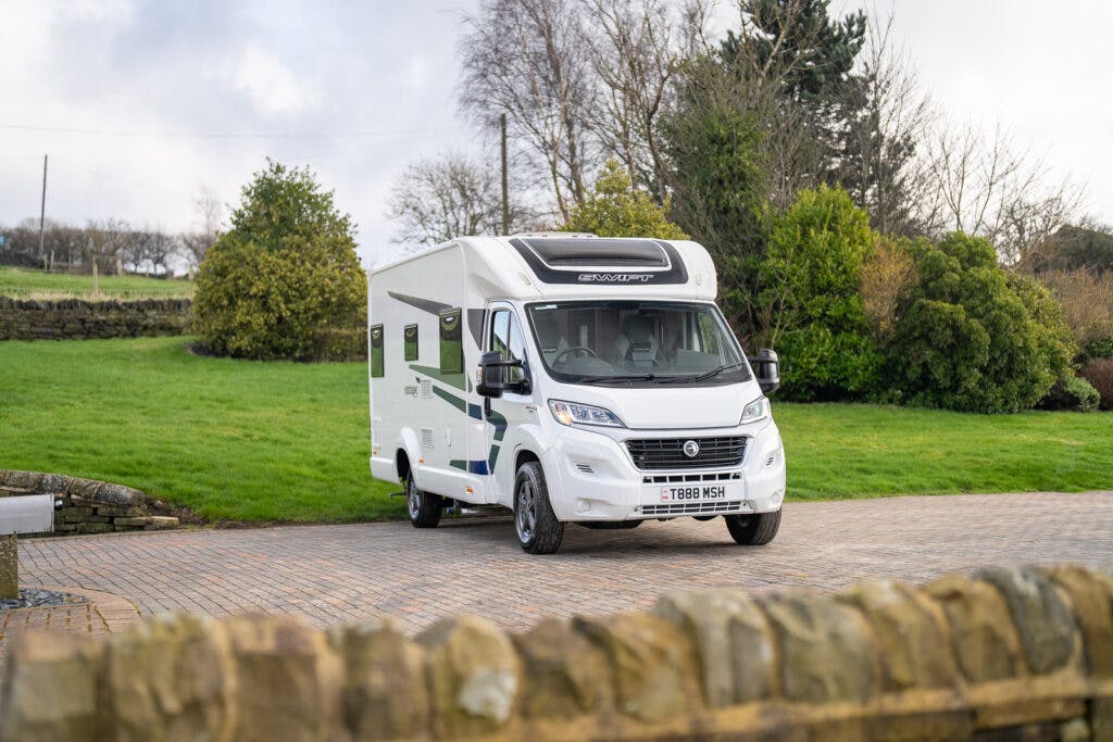 A white 2017 Swift Escape 664 RV is parked on a cobblestone driveway with a grassy area and trees in the background. The vehicle's license plate reads "T888 WSH." The sky is partly cloudy, and the surroundings are calm.