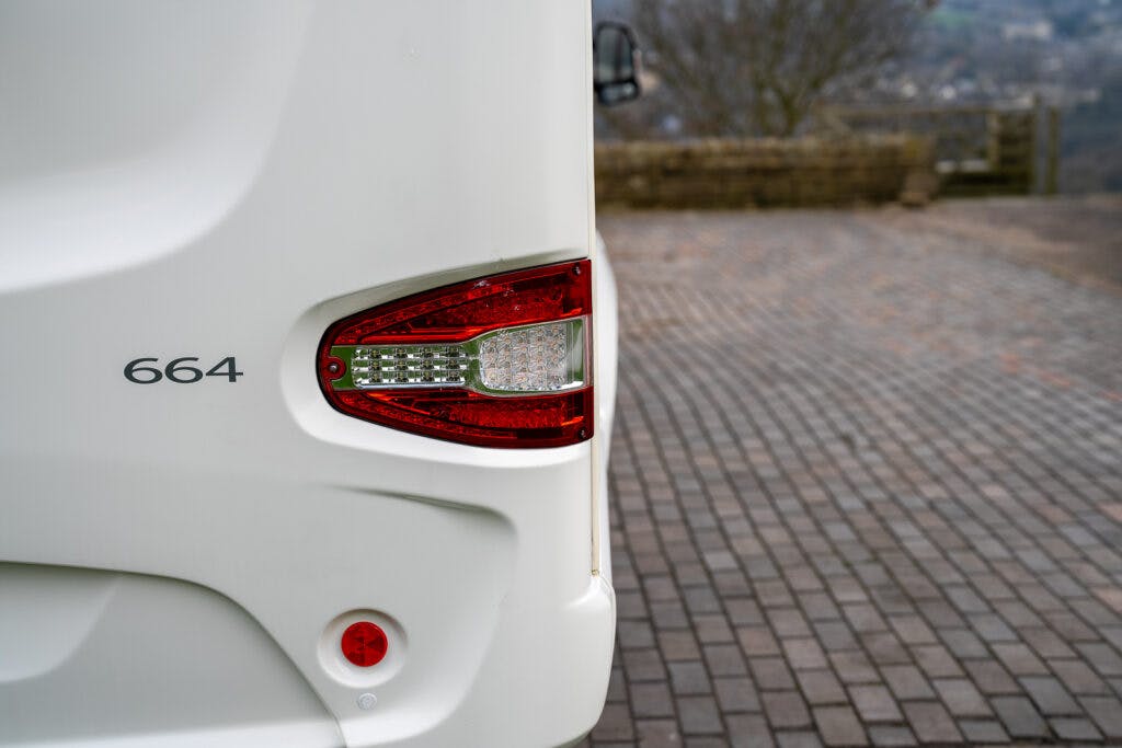 Close-up of the rear corner of a white 2017 Swift Escape 664. It features a tail light, a red circular reflector, and the number 664. The background includes a paved surface and an out-of-focus view of a landscape with trees and distant scenery.